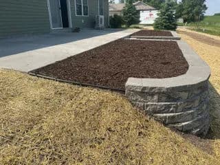 Mulched garden beds with stone edging beside a house on a sunny day.