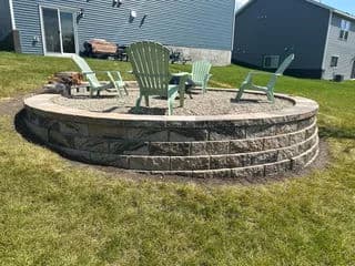 Outdoor fire pit area with stone seating and green Adirondack chairs in a backyard setting.