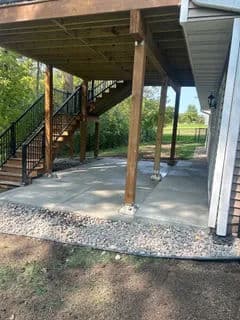 Underneath a wooden deck with stairs, featuring a concrete patio and landscaped area.
