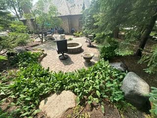 Outdoor garden scene featuring a circular stone patio, fire pit, and lush greenery.
